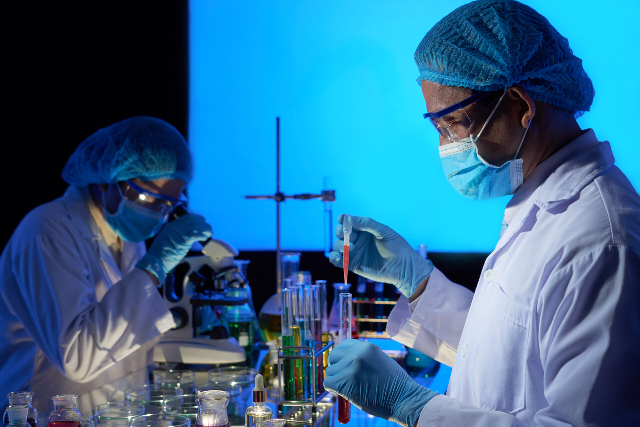 Gifted Asian scientist wearing rubber gloves and safety masks working together on creation of cancer vaccine, interior of dark laboratory on background